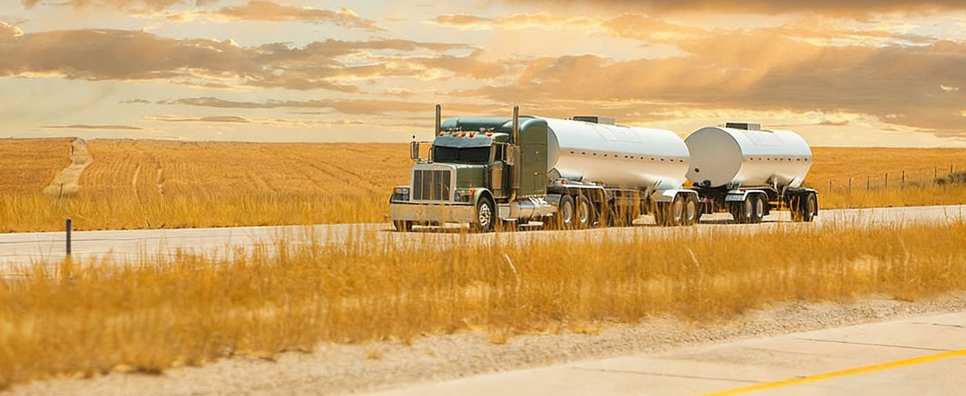 A heavy-duty silver tanker truck driving on a wide highway at sunset under a dramatic sky, representing long-haul transport and logistics.