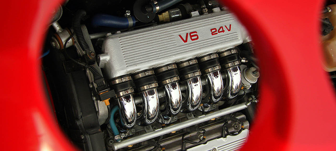 Close-up view of a V6 24-valve engine through a red circular opening, showing the valve cover, chrome intake manifolds, and associated components.